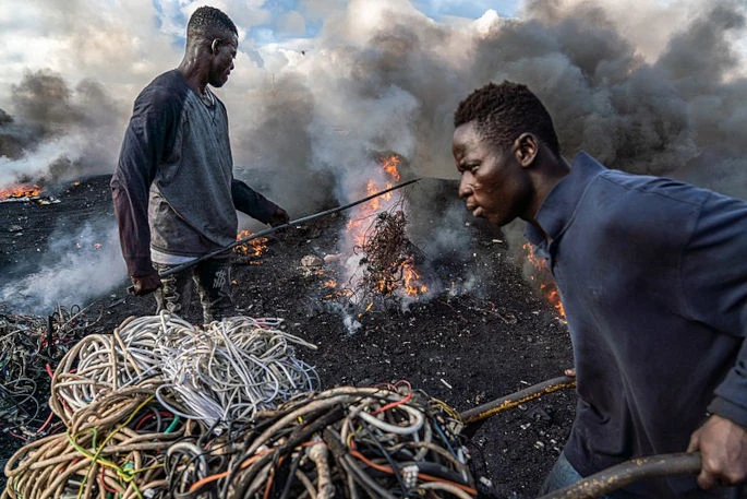 A young man is pictured burning electrical wires to recover copper at Agbogbloshie, Ghana, as another metal scrap worker arrives with more wires to be burned. A 2018 article in the &quot;International Journal of Cancer&quot; reports a <a href="https://onlinelibrary.wiley.com/doi/abs/10.1002/ijc.31902">correlation</a> between proximity to e-waste burn sites and childhood lymphoma. (Image by Muntaka Chasant, published under a <a href="https://spdx.org/licenses/CC-BY-SA-4.0.html">CC-BY-SA-4.0</a> license.)